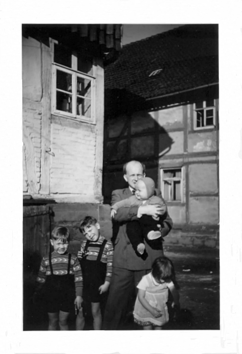 Schwarz-Weiß-Foto: Mein Vater mit mir im Arm. Auf dem Foto links von uns und vor uns drei Kinder von Nachbarn. Im Bildhintergrund rechts: Der Bauernhof der Familie Semmelroth. In Angerstein im Jahre 1954.